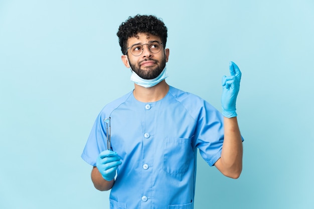 Foto dentista marroquí hombre sujetando herramientas aisladas en la pared azul con los dedos cruzando y deseando lo mejor
