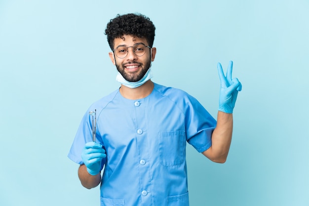Dentista marroquí hombre sujetando herramientas aisladas sonriendo y mostrando el signo de la victoria