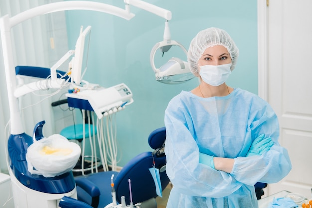 Una dentista con una máscara médica y guantes de goma posa para la cámara y cruza los brazos en su oficina.