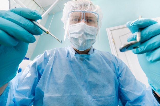Un dentista con una máscara protectora se sienta cerca y sostiene instrumentos en sus manos antes de tratar a un paciente en el consultorio dental