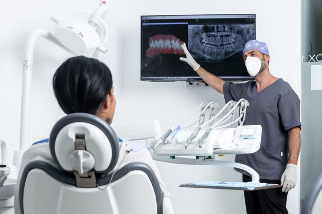 Dentista masculino apontando para um raio-x na frente de um patinet feminino sentado em uma cadeira em uma sala de clínica