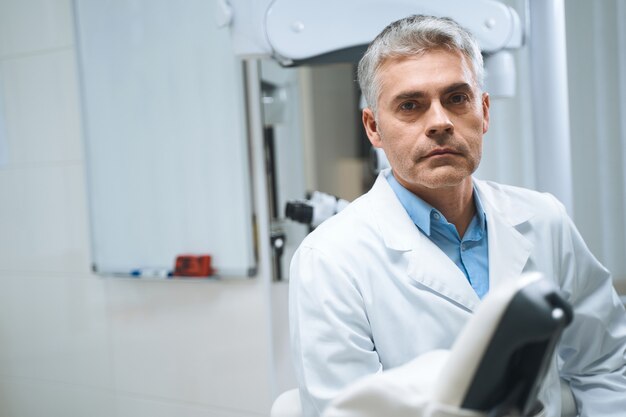 Dentista masculino em uniforme branco está de pé na clínica moderna e esperando a consulta com os pacientes.