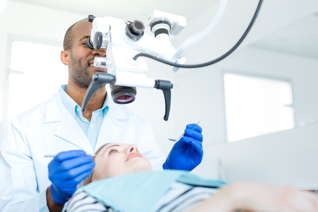 Dentista masculino guapo realizando un chequeo de los dientes de sus pacientes y usando un microscopio profesional para ese propósito
