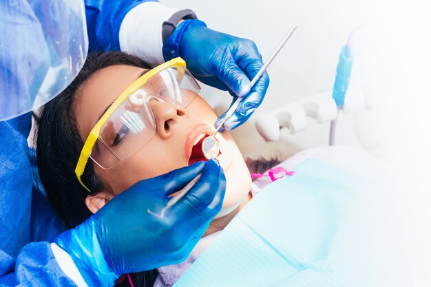 Foto dentista masculino latinx con traje de bioseguridad asistiendo y realizando un examen oral a una paciente