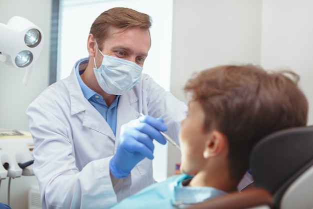 Dentista masculino maduro con máscara médica y guantes, examinando los dientes de un niño pequeño