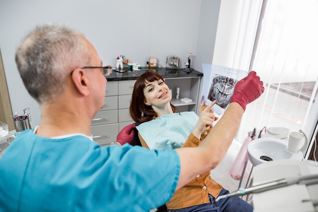 Foto dentista masculino senior en consultorio dental hablando con paciente femenino y preparándose para el tratamiento, mientras sostiene la imagen de rayos x de los dientes. mujer en clínica dental mirando la imagen de rayos con su dentista