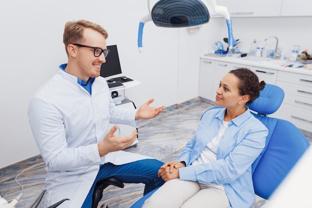 Foto dentista masculino sorridente explicando ao cliente sobre o procedimento odontológico na clínica com equipamentos modernos