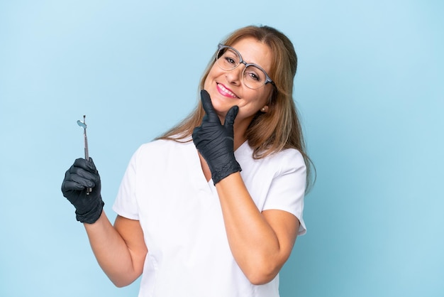 Dentista de mediana edad sosteniendo herramientas sobre fondo azul aislado feliz y sonriente