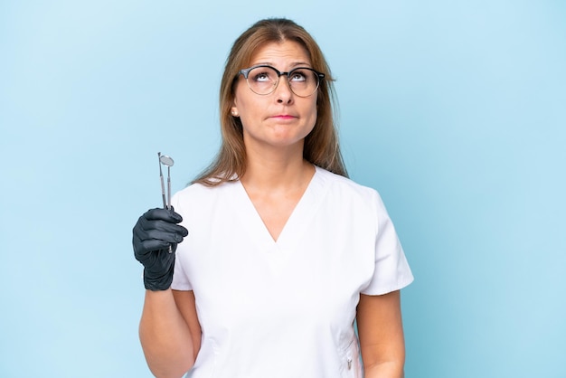 Dentista de mediana edad sosteniendo herramientas sobre fondo azul aislado y mirando hacia arriba