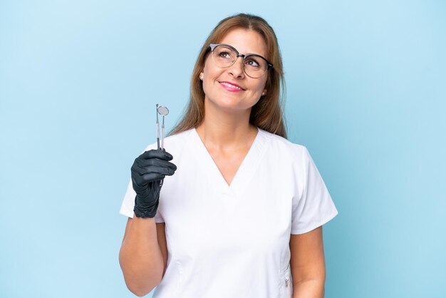 Dentista de mediana edad sosteniendo herramientas sobre un fondo azul aislado pensando en una idea mientras miraba hacia arriba