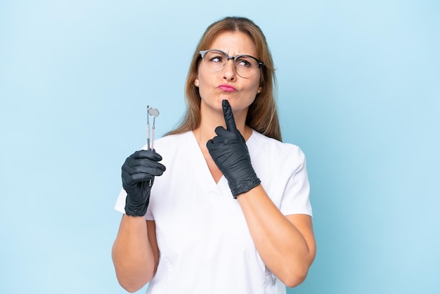 Dentista de mediana edad sosteniendo herramientas sobre un fondo azul aislado que tiene dudas mientras mira hacia arriba
