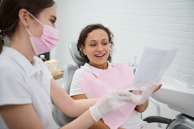 Dentista mostrando la mandíbula y los dientes en una radiografía y discutiendo con el paciente durante un examen dental, explicando los problemas de tratamiento de las consultas, en la odontología moderna.