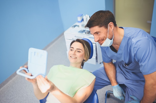 El dentista muestra a la paciente feliz los resultados del tratamiento con un espejo en el consultorio dental