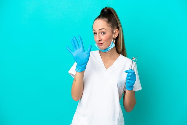 Dentista mujer caucásica sosteniendo herramientas aisladas sobre fondo azul contando cinco con los dedos