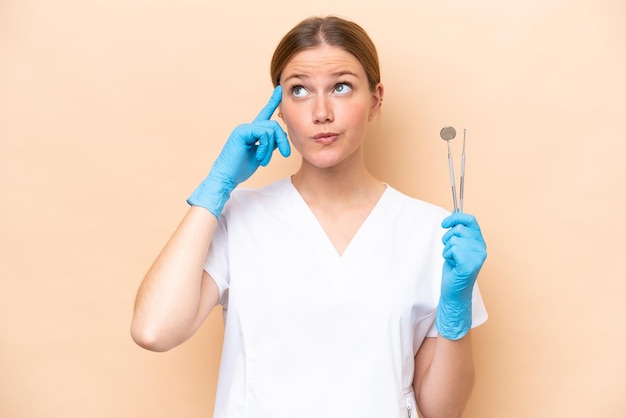 Dentista mujer caucásica sosteniendo herramientas aisladas sobre fondo beige con dudas y pensamiento