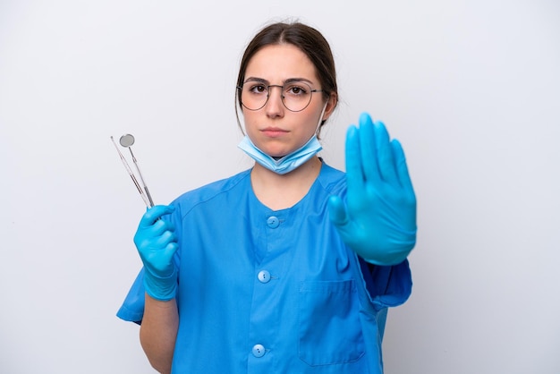 Dentista mujer caucásica sosteniendo herramientas aisladas sobre fondo blanco haciendo gesto de parada