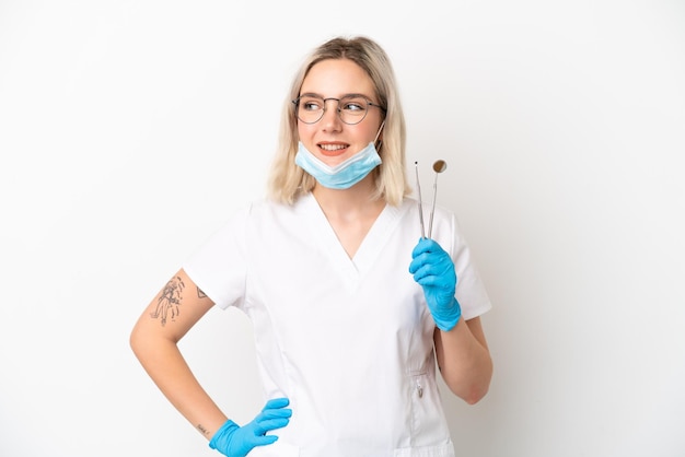 Dentista mujer caucásica sosteniendo herramientas aisladas sobre fondo blanco posando con los brazos en la cadera y sonriendo