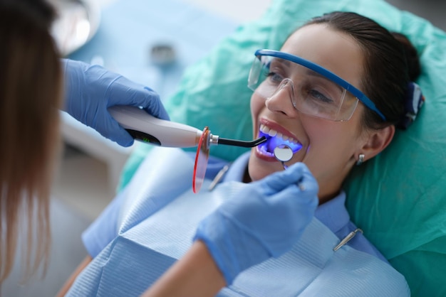 Dentista de la mujer joven en los guantes protectores que controlan el sello dental que fija la lámpara del fotopolímero durante el estomat ...