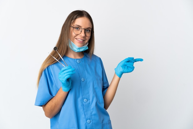 Dentista mujer lituana sosteniendo herramientas sobre pared aislada apuntando con el dedo hacia el lado