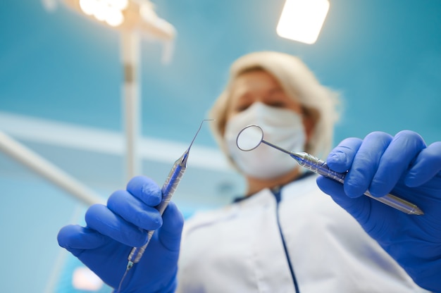 Dentista de mujer madura en máscara protectora y guantes se inclina hacia adelante mientras realiza un chequeo dental en el consultorio dental