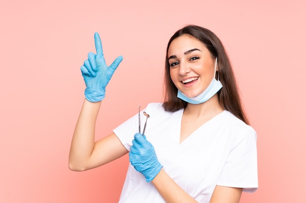 Dentista mujer sosteniendo herramientas aisladas en pared rosa apuntando con el dedo índice una gran idea