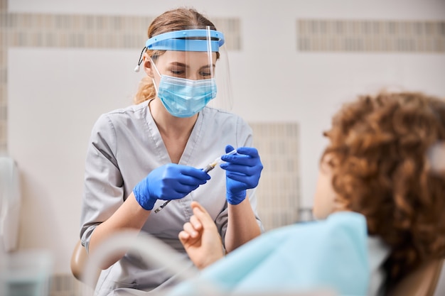 Dentista mujer sosteniendo una jeringa y preparándose para administrar anestesia al niño en un sillón dental