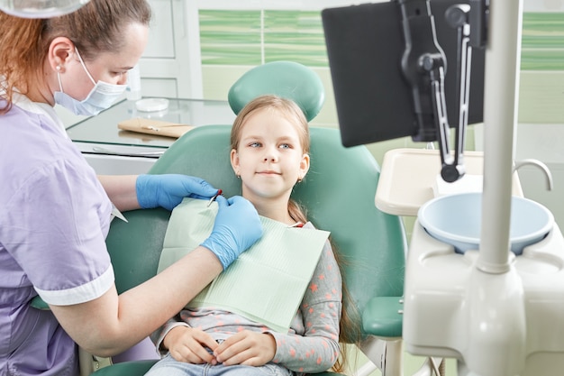 Dentista na máscara protetora e criança com tela no consultório de estomatologia. Jovem com na primeira consulta odontológica