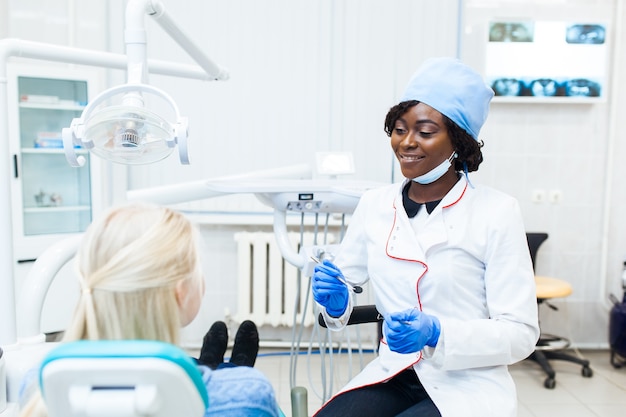 Dentista negro feminino no consultório odontológico, conversando com uma paciente do sexo feminino e se preparando para o tratamento. Equipamento médico moderno