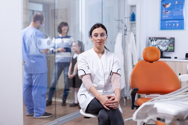 Dentista no consultório dentário, sentado na cadeira, enquanto o assistente está conversando com a mãe e a filha dela. Stomatolog na clínica de dentes professioanl sorrindo vestindo uniforme, olhando para a câmera.