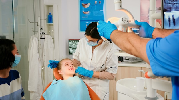 Dentista revisando con espejo la salud dental del paciente niña. Enfermera encendiendo la lámpara, médico hablando con un niño sentado en una silla estomatológica, preparándose para limpiar los dientes, tratando la masa afectada