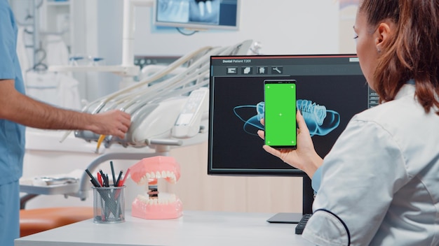 Dentista segurando verticalmente o smartphone com tela verde enquanto olha para os dentes digitalizados no computador no escritório. Especialista em cuidados dentários usando telefone celular com chroma key para fundo isolado