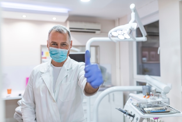 Dentista de sexo masculino envejecido medio feliz en la capa blanca que muestra los pulgares para arriba en la oficina dental de la clínica.