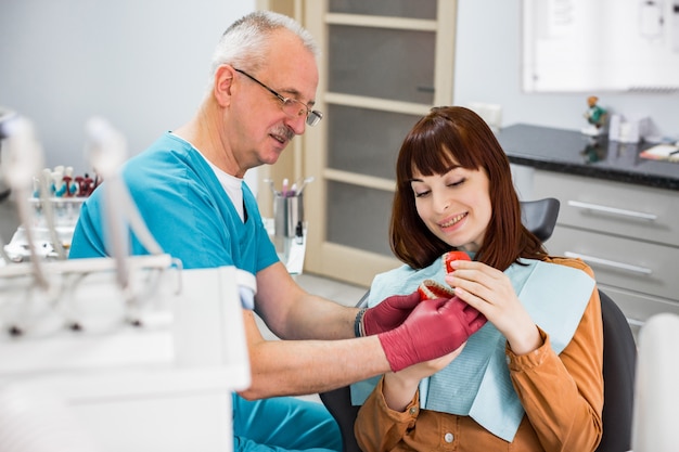 Dentista de sexo masculino mayor que muestra a la mujer bonita joven cómo cepillarse los dientes. Apuesto dentista senior hombre mostrando mandíbulas artificiales para su paciente en el consultorio dental