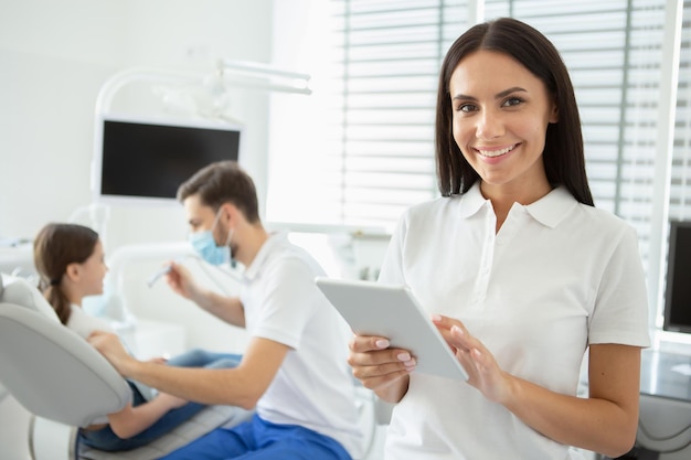 Dentista sorridente trabalhando em tablet digital e olhando para a câmera com médico masculino trabalhando com cliente em segundo plano