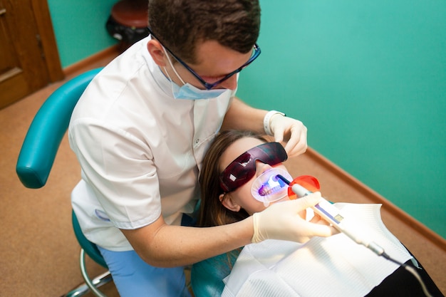 Dentista, usando a lâmpada ultravioleta de clareamento nos dentes. Paciente jovem na clínica odontológica encontra-se em óculos uv na cadeira do dentista