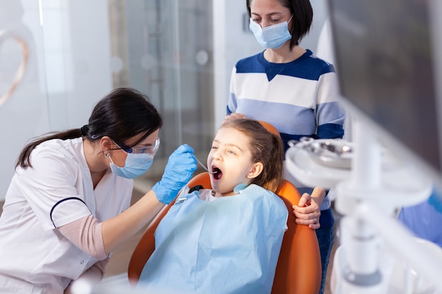 Dentista usando luvas durante o procedimento de cavidade na menina sentada na cadeira odontológica. Odontologista durante consulta de cárie infantil em consultório de estomatologia com tecnologia moderna.