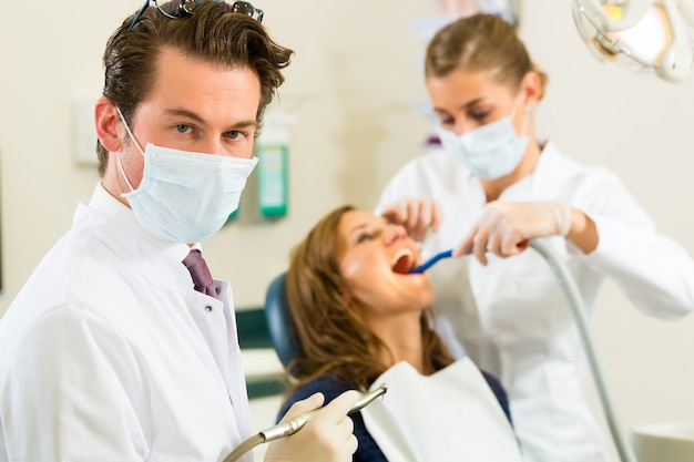 Dentistas, sua cirurgia, olhando para o espectador, em seu assistente, está dando tratamento a uma paciente do sexo feminino