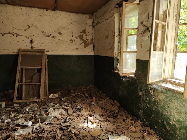 Dentro de una casa abandonada Basura esparcida por el suelo Grietas de pintura verde y telarañas en la pared La mitad superior blanca de la pared Ventanas abiertas a través de las cuales entra la luz del sol Marcos retirados