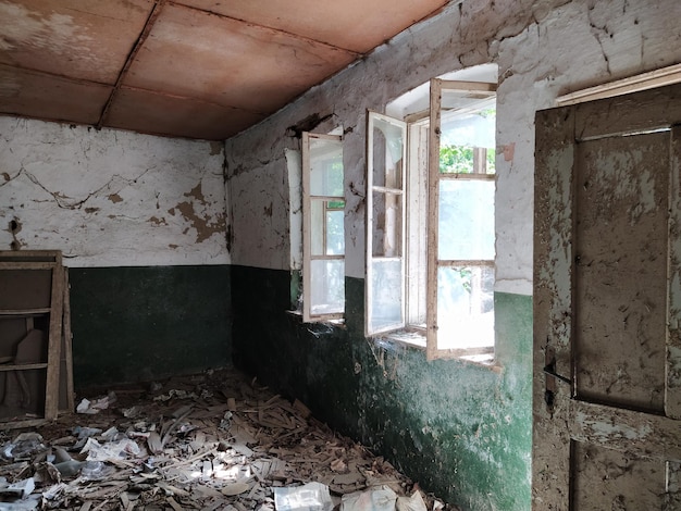 Dentro de uma casa abandonada Lixo espalhado no chão Tinta verde rachaduras e teias de aranha na parede A metade superior branca da parede Janelas abertas através das quais a luz do sol entra Porta velha