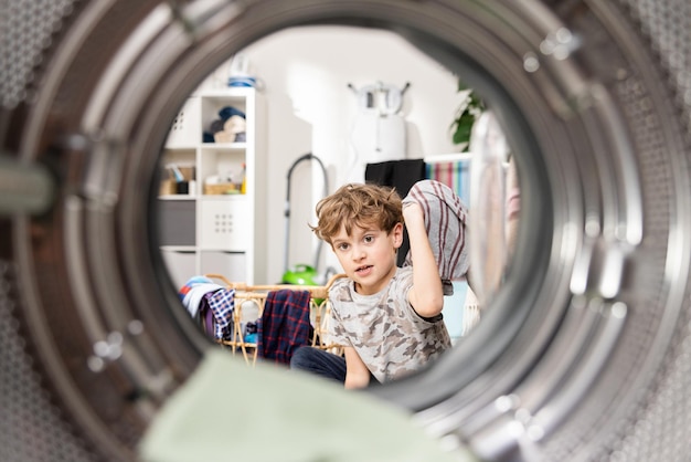 Dentro de la lavadora, un niño pequeño está poniendo ropa colorida en el tambor a través de la puerta abierta ayudando a su padre con las tareas domésticas