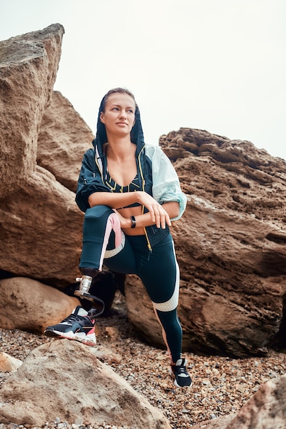 Depois do treino. Foto vertical de mulher atleta com deficiência em roupas esportivas com perna protética em pé na praia e olhando para a câmera. Conceito de esporte. Esporte lá fora. Desportista com deficiência.