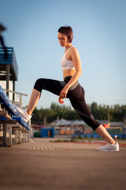 Deporte y bienestar. Chica fitness en zapatillas blancas haciendo ejercicios de estiramiento. Moda mujer deportiva con fuerte entrenamiento corporal musculoso. Colocar hembra estiramiento en el estadio al aire libre