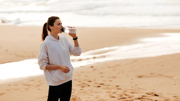 Deporte e hidratación Mujer joven en forma bebiendo agua mineral de una botella descansando después del entrenamiento junto al mar