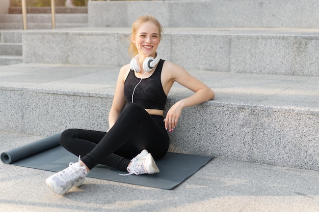 Deporte y Fitness Adulto joven Mujer caucásica Sentado Colchoneta de ejercicio Piso de concreto Descansar después de la mañana Entrenamiento al aire libre Verano Parque Atlético Mujer Tomar un descanso Escuchar música Auriculares