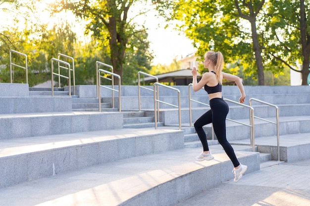 Deporte y Fitness. Ajuste mujer corriendo escaleras verano soleado mañana caucásico atlético femenino jogging al aire libre entrenamiento cardiovascular activo estilo de vida saludable vestido negro ropa deportiva motivación concepto