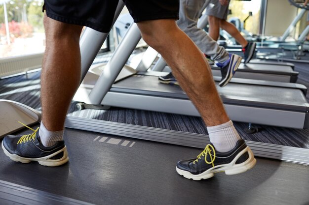 deporte, fitness, estilo de vida, tecnología y concepto de personas: cierre de las piernas de los hombres caminando en cintas de correr en el gimnasio