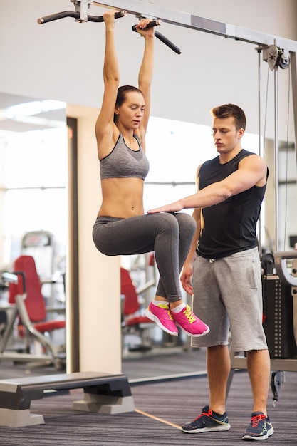 deporte, fitness, estilo de vida, trabajo en equipo y concepto de personas - mujer joven con entrenador colgando en la barra y haciendo levantamientos de piernas en el gimnasio