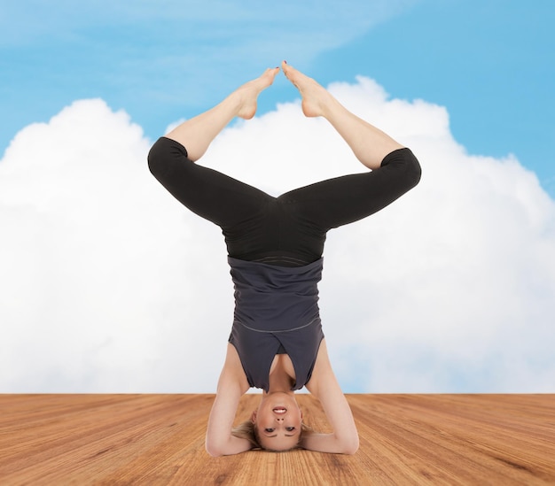 deporte, fitness, yoga, gente y concepto de salud - mujer joven feliz haciendo ejercicio de cabeza en una litera de madera sobre nubes blancas y fondo de cielo azul