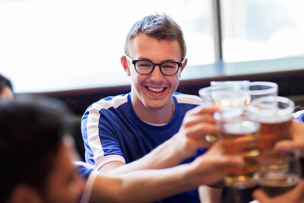 deporte, fútbol, gente y concepto de ocio - amigos felices o aficionados al fútbol que tintinean vasos de cerveza en el bar o pub
