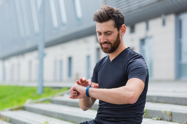 Deporte hombre caucásico mirando reloj inteligente en corredor de pista moderno usando usar pulsera de fitness smartwatch Retrato en forma atleta masculino comprobar resultado trotar en ropa deportiva en la calle parque urbano de la ciudad al aire libre